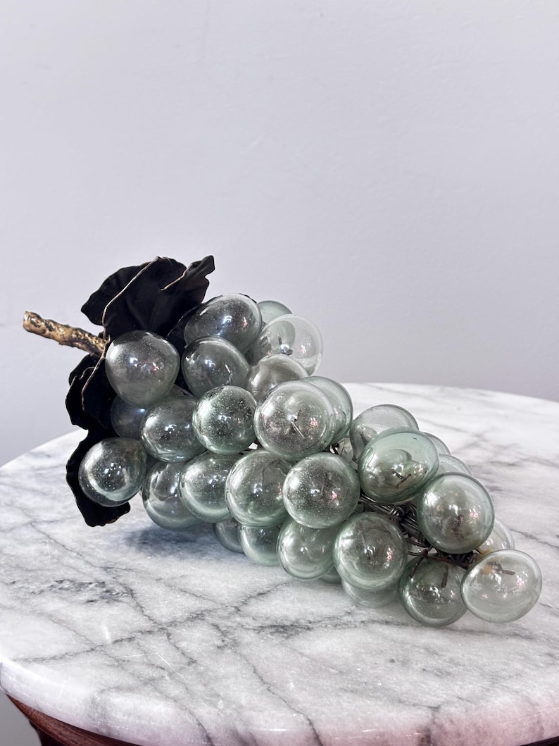 May include: A decorative cluster of translucent, pale green glass grapes attached to a dark, textured metal stem and leaf. The individual glass spheres are rounded and smooth, creating a realistic bunch of fruit with a metallic accent at the top.