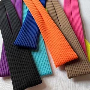 May include: A selection of vibrantly coloured, ribbed knit fabric strips. The palette includes black, purple, blue, teal, orange, brown, pink, and yellow. The strips are arranged in a fan shape, highlighting the diverse colours and textures of the fabric.