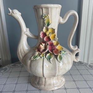 Könnte beinhalten: Cremefarbener Keramikkrug mit schwanenförmigem Ausguss und gebogenem Griff. Der Krug ist mit farbenfrohen, dreidimensionalen floralen Akzenten in Rosa, Gelb und Blau verziert. Der Sockel hat ein Podestdesign.