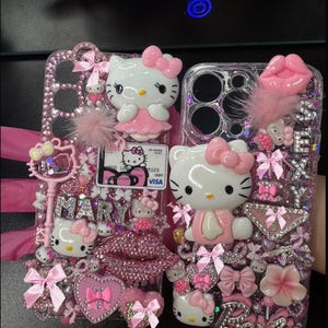 K&ouml;nnte beinhalten: Zwei verzierte Handyh&uuml;llen mit Hello Kitty-Figuren, rosa Schleifen und glitzernden Strasssteinen. Eine H&uuml;lle hat den Namen "MARY" in silbernen Buchstaben, die andere "Barbie". Beide H&uuml;llen haben eine Visa-Karte.
