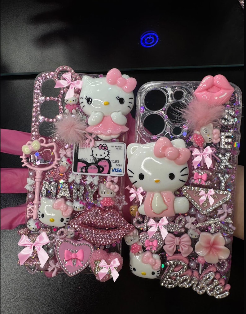 K&ouml;nnte beinhalten: Zwei verzierte Handyh&uuml;llen mit Hello Kitty-Figuren, rosa Schleifen und glitzernden Strasssteinen. Eine H&uuml;lle hat den Namen "MARY" in silbernen Buchstaben, die andere "Barbie". Beide H&uuml;llen haben eine Visa-Karte.