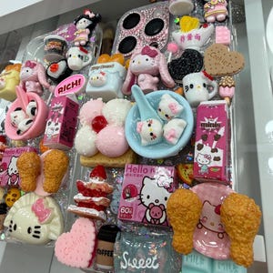 Pu&ograve; includere: Una custodia trasparente per telefono decorata con vari ciondoli e miniature di cibo a tema Hello Kitty. La custodia presenta elementi rosa, bianchi e blu, tra cui un biscotto a forma di cuore, pollo fritto e una tazza di caff&egrave;. La parola "Sweet" &egrave; visibile.
