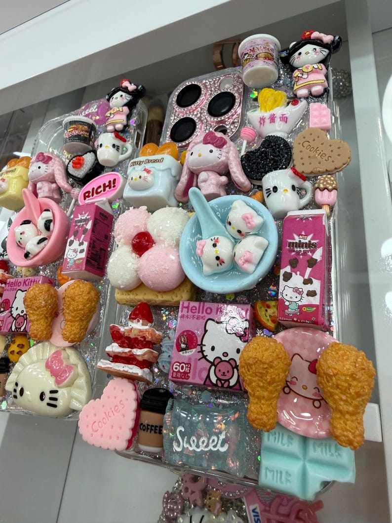Pu&ograve; includere: Una custodia trasparente per telefono decorata con vari ciondoli e miniature di cibo a tema Hello Kitty. La custodia presenta elementi rosa, bianchi e blu, tra cui un biscotto a forma di cuore, pollo fritto e una tazza di caff&egrave;. La parola "Sweet" &egrave; visibile.