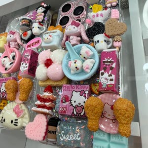 Pu&ograve; includere: Una custodia trasparente per telefono decorata con vari ciondoli in miniatura di cibo e personaggi. I ciondoli includono personaggi di Hello Kitty, biscotti, pollo fritto e cibi in miniatura. La custodia ha uno sfondo glitterato e la parola "Sweet" &egrave; visibile.