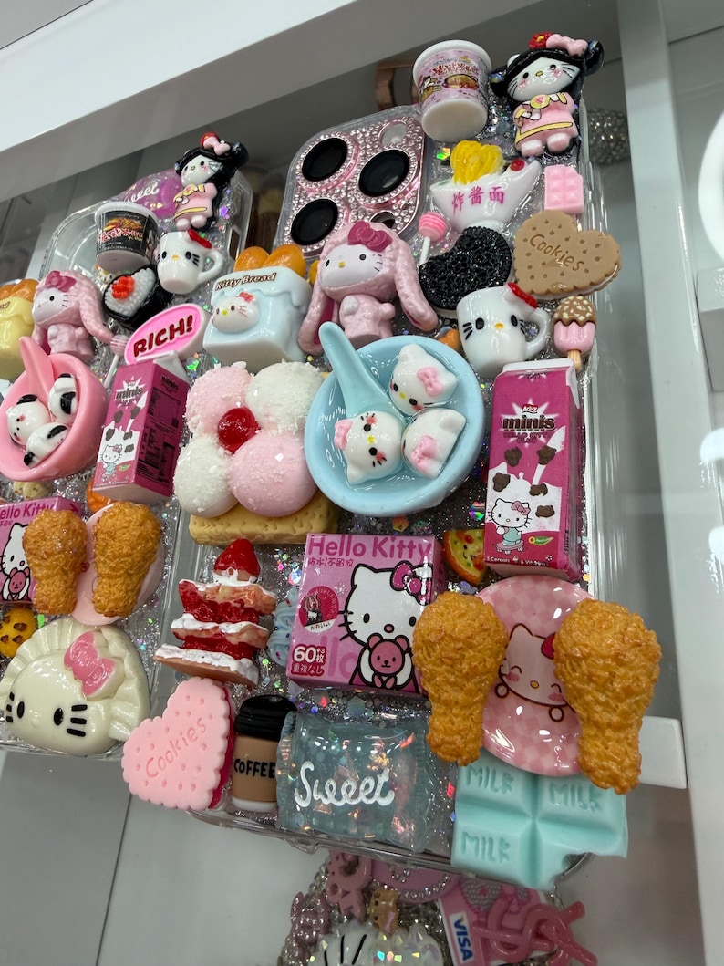 Pu&ograve; includere: Una custodia trasparente per telefono decorata con vari ciondoli in miniatura di cibo e personaggi. I ciondoli includono personaggi di Hello Kitty, biscotti, pollo fritto e cibi in miniatura. La custodia ha uno sfondo glitterato e la parola "Sweet" &egrave; visibile.