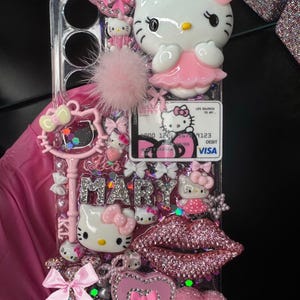 K&ouml;nnte beinhalten: Eine transparente Handyh&uuml;lle, verziert mit rosa Hello Kitty Figuren, Schleifen und dem Namen "MARY" in silbernen Strasssteinen. Die H&uuml;lle hat auch einen rosa Pompon, ein Herz und ein glitzerndes Lippen-Design.
