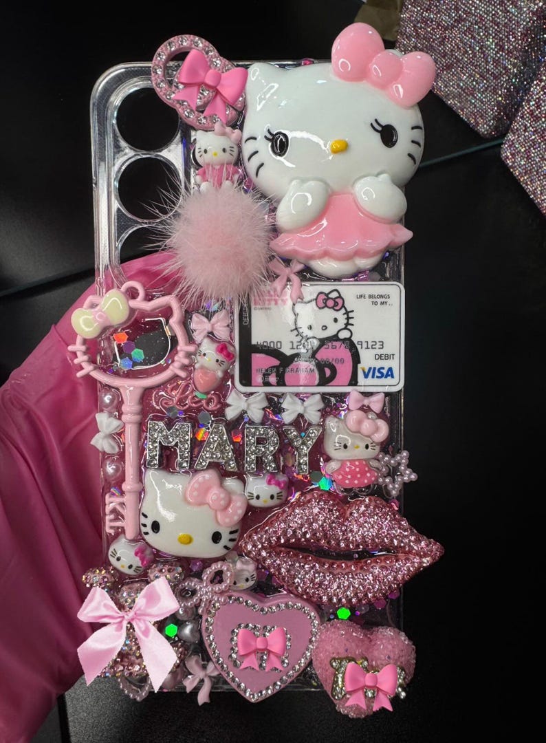 K&ouml;nnte beinhalten: Eine transparente Handyh&uuml;lle, verziert mit rosa Hello Kitty Figuren, Schleifen und dem Namen "MARY" in silbernen Strasssteinen. Die H&uuml;lle hat auch einen rosa Pompon, ein Herz und ein glitzerndes Lippen-Design.