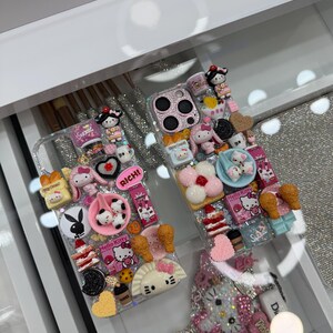 Pu&ograve; includere: Due custodie trasparenti per telefono decorate con vari ciondoli, tra cui personaggi di Hello Kitty, miniature di cibo e altri elementi decorativi. Una custodia presenta la scritta "RICH!". Le custodie sono esposte in un cassetto bianco.