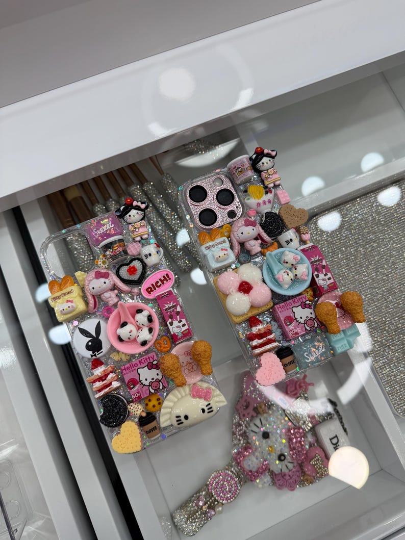 Pu&ograve; includere: Due custodie trasparenti per telefono decorate con vari ciondoli, tra cui personaggi di Hello Kitty, miniature di cibo e altri elementi decorativi. Una custodia presenta la scritta "RICH!". Le custodie sono esposte in un cassetto bianco.
