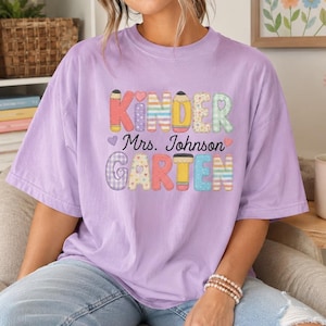 May include: A light purple short-sleeve t-shirt featuring the text "KINDER GARTEN" in colorful, patterned block letters with pencil accents and the name "Mrs. Johnson" printed in black script in the center of the graphic design.