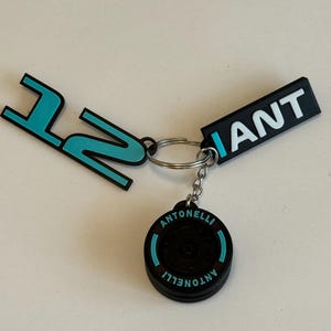 Könnte beinhalten: Ein Schlüsselanhänger mit einem schwarz-türkisen Design. Der Schlüsselanhänger enthält das Wort "ANT" in Weiß auf einem schwarzen Rechteck, eine türkisfarbene "12"-Form und einen runden Anhänger mit "ANTONELLI"-Aufdruck.