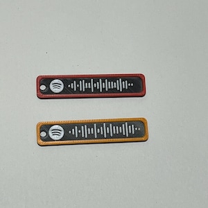 Könnte beinhalten: Zwei rechteckige Spotify-Code-Tags. Eines ist rot mit schwarzem Zentrum und weißem Spotify-Logo. Das andere ist orange mit schwarzem Zentrum und weißem Spotify-Logo. Beide haben eine Reihe vertikaler Linien.