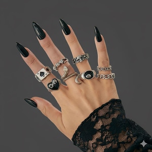 May include: A hand displaying several silver rings with diverse motifs, such as a snake, a heart, and a playing card. The hand features long, black fingernails and is complemented by a black lace sleeve.