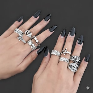 May include: A selection of silver rings featuring diverse designs such as stars, geometric patterns, and floral details. The rings are shown on two hands with long, black polished nails, set against a dark backdrop.