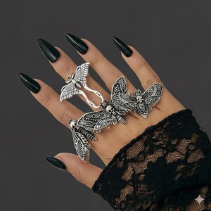 May include: Silver-toned moth rings with intricate details, including skull accents, are displayed on a hand with long, dark nails. The rings feature various moth designs, showcasing a gothic or alternative aesthetic. The hand is adorned with a black lace sleeve.
