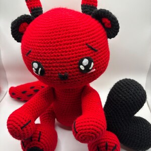 May include: A handmade red crochet plush toy with black accents. The toy has large, expressive eyes, small horns, and a black tail. The plush is sitting on a white surface. The toy's design is reminiscent of a cartoon character.