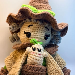 May include: A handmade crochet doll with a whimsical design. The doll features a tan face, brown hair, and a pointed brown hat with green accents. It wears a tan and brown outfit with a pocket holding a small, white creature with a green top.
