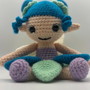 May include: A handmade crochet doll in the shape of a fairy. The doll has a light skin tone, with blue hair and accents, and large, shiny black eyes. The fairy is wearing a lavender and green outfit and has small wings.