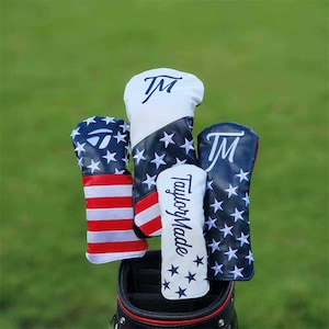 May include: A collection of golf club head covers with patriotic designs. The covers feature white, navy blue, and red colors with star and stripe patterns. Some covers have the TaylorMade logo and the initials "TM".