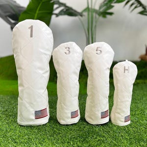 May include: Set of four white golf club head covers. Each cover is quilted and features a number or letter in silver. The covers have an American flag patch at the bottom. The numbers are 1, 3, and 5, and the letter is H.