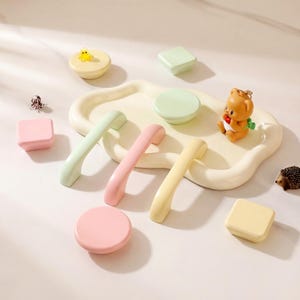 May include: A collection of pastel-colored cabinet hardware, including handles and knobs, arranged on a cream-colored tray. The hardware is in shades of pink, green, and yellow. Decorative elements include a small bear figurine and a hedgehog.