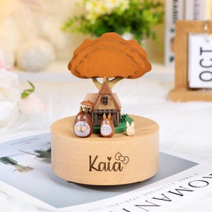 May include: A wooden music box featuring a miniature scene with a house, tree, and cartoon characters. The box is light brown with the name "Kaia" printed on the front. The tree has a brown, layered design, and the characters are brown and white.