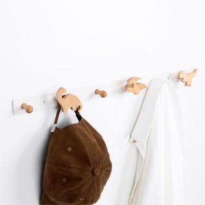 May include: A white wall-mounted coat rack with wooden dinosaur-shaped hooks. A brown baseball cap and a white shirt hang from the hooks. The rack has a simple design, perfect for a child's room or nursery.
