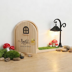Pode incluir: Uma porta de fadas de madeira com uma pequena janela e uma aldrava dourada. A porta tem a palavra "Love" e os nomes "Amily & Ethan". Um poste de luz preto com uma lâmpada acesa e decorações de cogumelos vermelhos estão ao lado da porta.