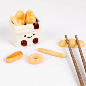 May include: A charming ceramic chopstick rest set featuring bread shapes. The set includes a smiling white container with bread inside, plus a baguette, a donut, and a bread roll. Wooden chopsticks are also included.
