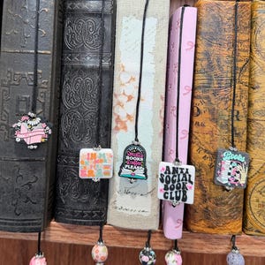 May include: A collection of vintage books with decorative bookmarks. The bookmarks have various designs and text, including "If my book is open your mouth is closed", "Anti Social Book Club", and "A book a day". The books are in various colors and sizes.