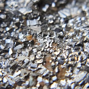 May include: A macro view of a mineral specimen featuring a dense cluster of small, metallic, silver-gray crystals with sharp, angular facets. The crystalline structure displays a reflective, high-gloss finish with subtle hints of brown earth tones.