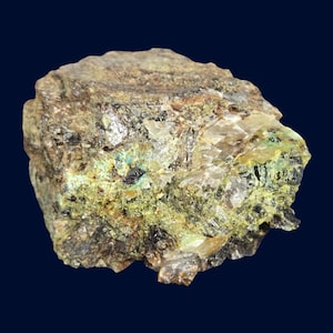 Op de afbeelding: Een close-up van een ruw, onregelmatig gevormd mineraalmonster. De steen heeft een mix van kleuren, waaronder olijfgroen, bruin en zwart, met kristallijne structuren en een licht reflecterend oppervlak. De achtergrond is donkerblauw.
