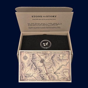 May include: An open cardboard box displaying a small, clear, circular container with several small, light-colored stones. Below the container is a map of the Virginia City Mining District. The box has the text "STONE TO STORY".