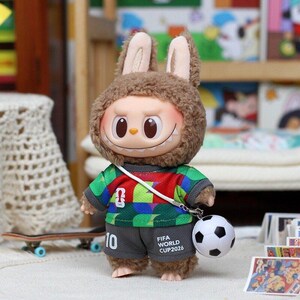 May include: A plush rabbit figure wearing a green, red, and blue patterned soccer jersey and gray shorts with the text FIFA WORLD CUP 2026 and the number 10. The figure has a white strap across its chest attached to a small black and white soccer ball.