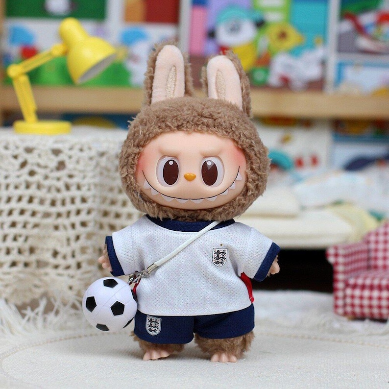 May include: A plush toy with brown fur and long ears wearing a white and navy blue soccer jersey and shorts set. The jersey features a small emblem on the chest, and the shorts have a matching logo. A small black and white soccer ball is attached.