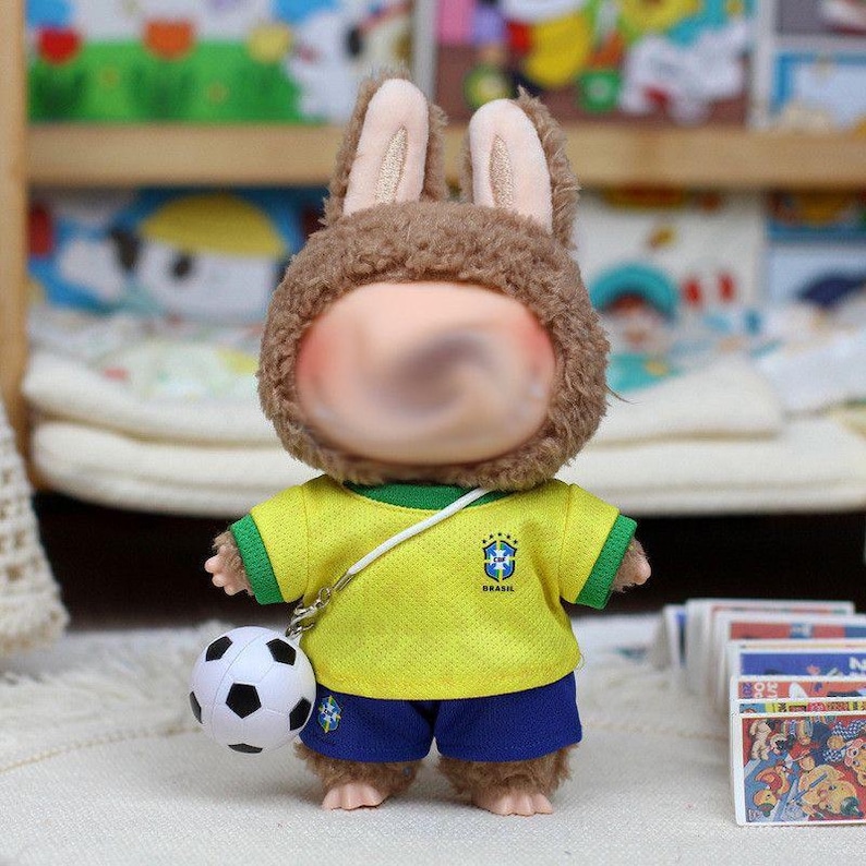 May include: A plush brown rabbit doll wearing a yellow Brazil soccer jersey with green trim and blue shorts. The doll has a white strap across its chest attached to a small black and white soccer ball. The jersey features the Brazil crest.