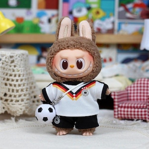 May include: A small doll with brown fuzzy rabbit ears and a wide, toothy grin. The doll wears a white soccer jersey featuring a black, red, and gold chevron pattern across the chest, a German national team crest, and black shorts with a matching crest.