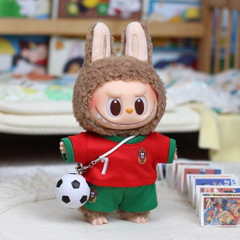 May include: A plush toy figure with a brown fuzzy rabbit hood, large eyes, and a wide grin. The figure wears a red soccer jersey with green trim, the number 7, and a crest, paired with green shorts. A small white and black soccer ball is attached.