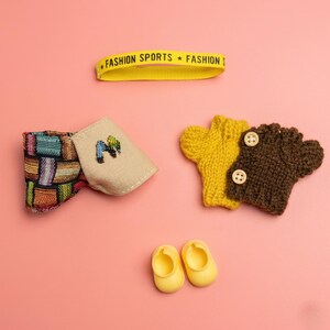 Puede incluir: Colecci&oacute;n de accesorios para beb&eacute;s sobre un fondo rosa. Incluye una diadema amarilla con texto, una pajarita colorida, una bolsa beige con bordado, dos guantes de punto y dos zapatos de beb&eacute; amarillos.