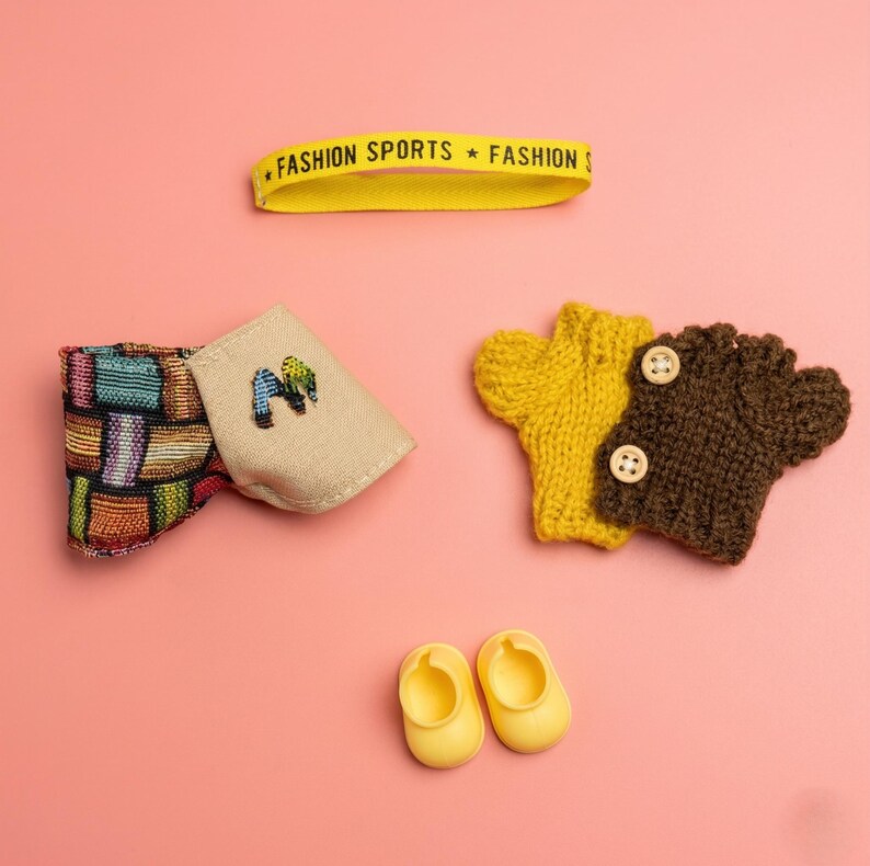 Puede incluir: Colecci&oacute;n de accesorios para beb&eacute;s sobre un fondo rosa. Incluye una diadema amarilla con texto, una pajarita colorida, una bolsa beige con bordado, dos guantes de punto y dos zapatos de beb&eacute; amarillos.