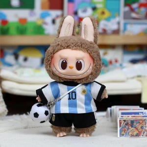 May include: A plush doll wearing a brown fuzzy rabbit hood and a light blue and white striped soccer jersey with the number 10 and an AFA logo on the chest. The doll wears black shorts and carries a small white and black soccer ball on a strap.