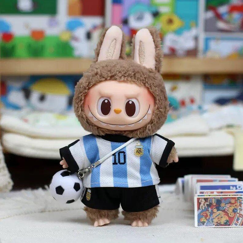 May include: A plush doll wearing a brown fuzzy rabbit hood and a light blue and white striped soccer jersey with the number 10 and an AFA logo on the chest. The doll wears black shorts and carries a small white and black soccer ball on a strap.