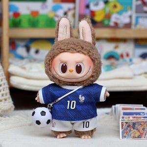 May include: A plush doll with brown fur ears wearing a blue soccer jersey with the number 10 and a French rooster emblem, paired with white shorts featuring the number 10. A small white and black soccer ball is attached by a white strap.