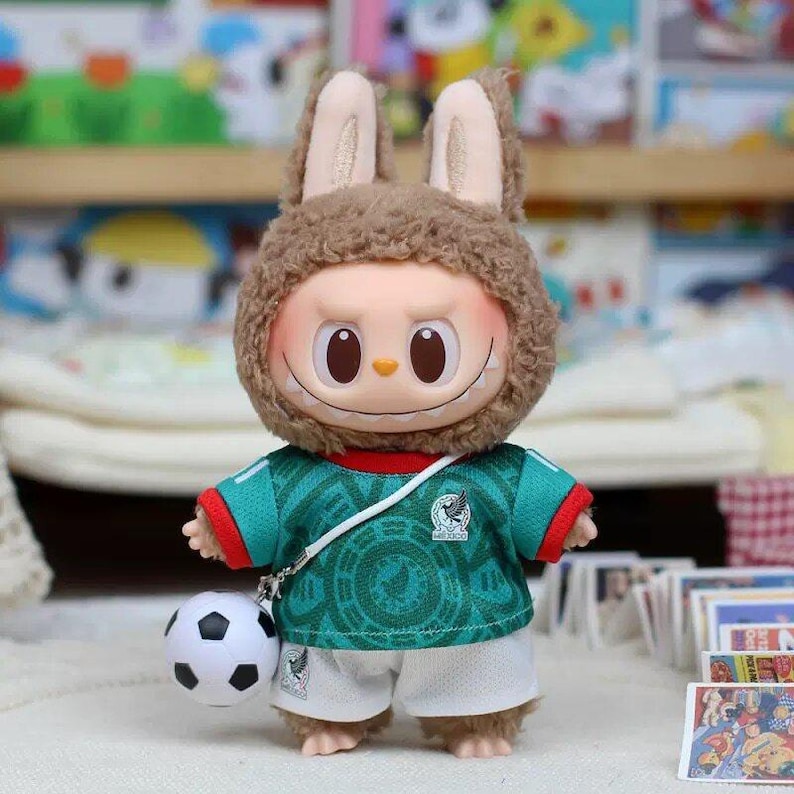 May include: A plush toy figure with brown fuzzy rabbit ears and a wide, toothy grin. The figure wears a green soccer jersey with red trim and white shorts, featuring a small soccer ball attached by a white strap across the chest.