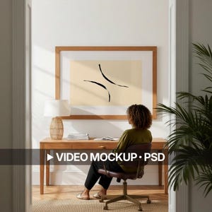 Peut inclure: Un bureau en bois avec une lampe et une œuvre d'art abstraite encadrée. Une personne est assise au bureau sur une chaise marron. L'œuvre présente des lignes courbes noires sur fond crème. Le texte "VIDEO MOCKUP • PSD" est visible.