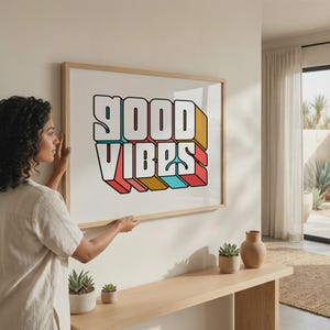 May include: A framed art print with the words "GOOD VIBES" in a 3D retro style. The text is white with black outlines and colorful shadows in red, yellow, and blue. The print is in a light wood frame.