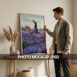 以下が含まれることがあります： 紫色の花畑を自転車で走る女性が描かれた額入りのアートプリント。 アートワークは木製のフレームに入っており、人が持っています。 下部には「PHOTO MOCKUP • PSD」というテキストがあります。