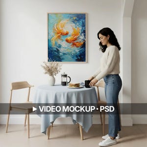 Pode incluir: Uma pintura emoldurada de dois peixes koi laranja nadando em uma cena de água azul e branca está pendurada acima de uma mesa montada para uma refeição. Uma mulher está colocando uma caneca preta na mesa. A mesa é coberta com uma toalha de mesa azul clara. O texto "VIDEO MOCKUP • PSD" está na parte inferior.