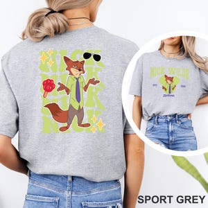 May include: Sport grey t-shirt featuring a cartoon fox character with sunglasses, a tie, and a lollipop. The design includes the text "Nick Wilde" and "Zootopia" in a green and yellow color scheme. The shirt is a casual wear item.