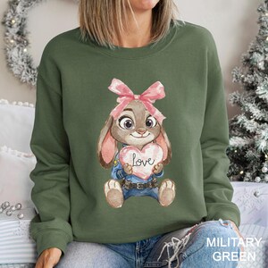 May include: Military green sweatshirt with a watercolour illustration of a bunny holding a heart-shaped sign with the word "Love". The bunny wears a pink bow and a police uniform. The sweatshirt is a solid military green colour.
